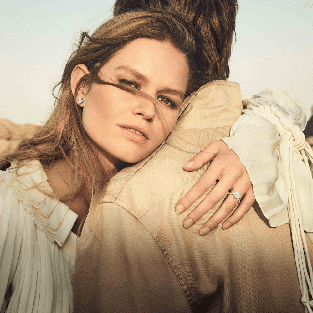 Image of Louis Vuitton photoshoot of woman wearing fringe beige jacket with diamond earrings and diamond rings holding man whose face isn't showing