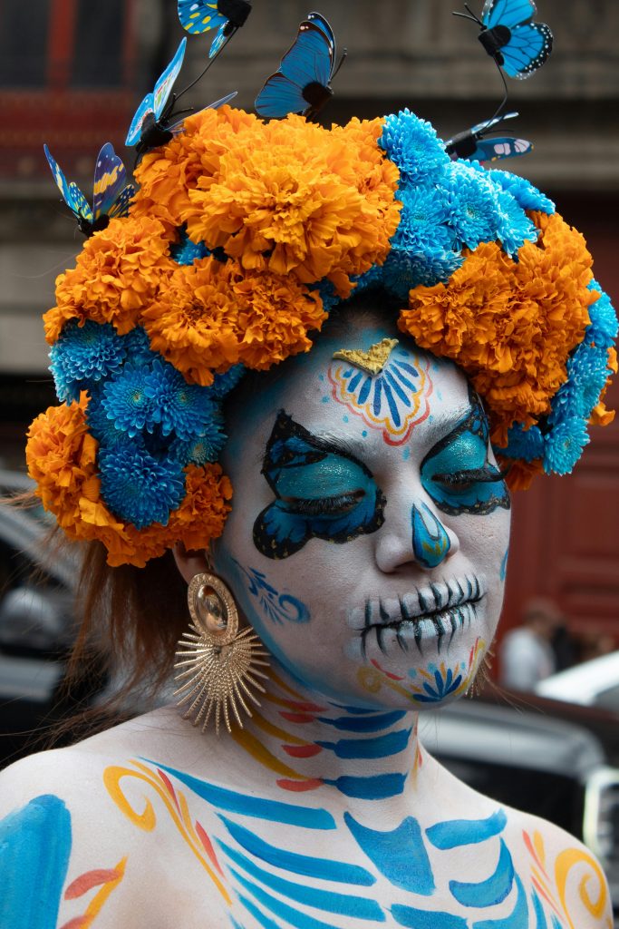 Image of woman wearing day of the dead skull makeup, blue and marigold flower crown, and large earrings